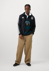 Μαύρο μπουφάν με φερμουάρ και λευκές ρίγες, πράσινο και ναυτικό μπλε πουκάμισο με ρίγες, μπεζ παντελόνι σε φαρδιά γραμμή και μαύρα αθλητικά. Περιέχει λογότυπα.