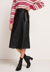 Falda envolvente de cuero negro con una textura suave, de longitud media hasta la pantorrilla y detalle de cintura atada, combinada con botas de tacón negro hasta el tobillo.