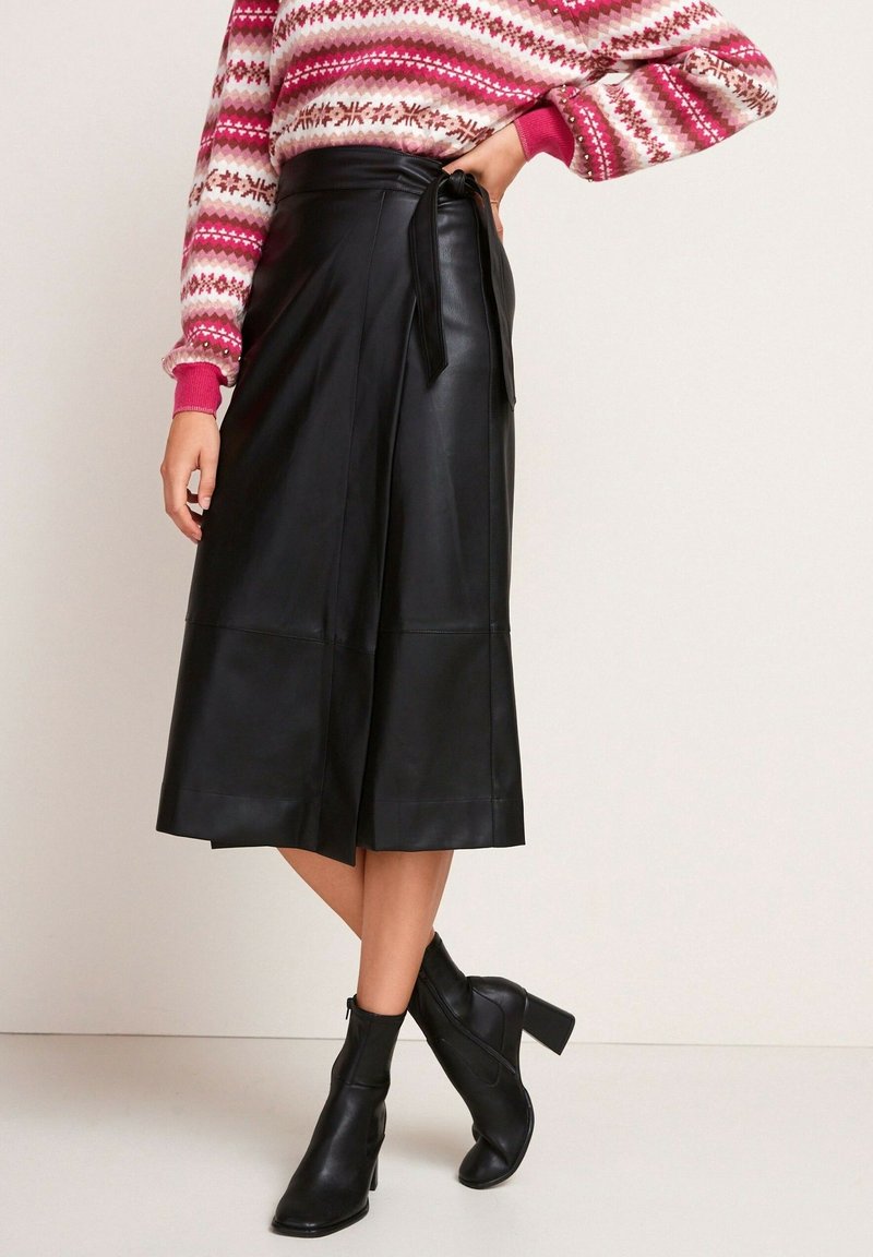 Falda envolvente de cuero negro con una textura suave, de longitud media hasta la pantorrilla y detalle de cintura atada, combinada con botas de tacón negro hasta el tobillo.