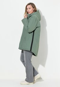 Ljusgrön huva jacka med svarta sidostråk, som har en avslappnad passform, stora fickor och en slät textur. Bärs över grå utsvängda jeans.