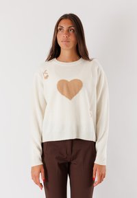 Donna con lunghi capelli castani, che indossa un maglione bianco con un grande cuore beige e pantaloni marroni, in piedi su uno sfondo semplice.