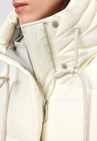 Närbild av en person som bär en krämfärgad vadderad jacka med en delvis dragkedjad hög krage och dragsnöre som är knutet i en rosett på huven.