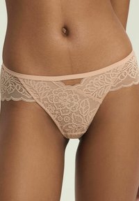 Koronkowy string w kolorze nude z kwiecistymi wzorami, z gładkim pasem w talii i ozdobnymi falbankami. Miękka tekstura materiału.
