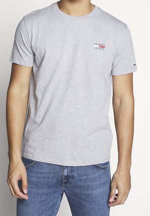 Mand iført lysegrå Tommy Jeans t-shirt med lille logo på brystet og mørkeblå jeans, stående mod en ensfarvet baggrund.