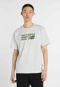 Camiseta de algodón gris con un estampado en el frente que dice "new balance" y "calzado deportivo" en verde oscuro y negro. Corte regular, cuello redondo.
