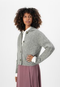Cardigan grigio a maglia con colletto, caratterizzato da grandi bottoni e polsini a coste. Indossato sopra una camicia bianca e una gonna rosa. Tessuto testurizzato.