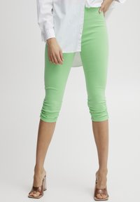 Zöld capri leggings rugalmas anyagból, fodros szegélyekkel. Fehér, gombos inggal és átlátszó sarkú szandállal párosítva.