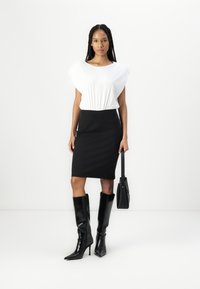 Haut blanc à manches courtes avec taille froncée, jupe crayon noire, bottes glossy noires jusqu'aux genoux et un sac à main noir.