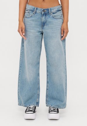 Jeans largos de lavagem azul clara, feitos de denim, com fecho de botão e ziper, cinco bolsos e punhos acabados. Usados com ténis pretos.