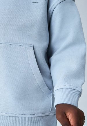 Nahaufnahme einer Person, die ein hellblaues Sweatshirt mit einer Fronttasche und gerippter Manschette trägt, wobei ein Teil des Arms und der Hand sichtbar ist.