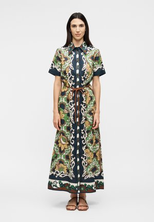 Γυναίκα στέκεται προς τα εμπρός φοράει ένα μακρύ, πουκαμισοφόρεμα με πράσινα, ναυτικά και καφέ ανθισμένα σχέδια, δεμένο με μια λεπτή καφέ ζώνη και σανδάλια.