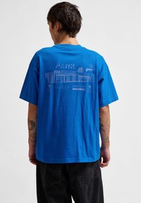 Blå bomullströja med grafiskt tryck på ryggen, som har en stadiondesign och vit textinformation. Kort ärm och avslappnad passform.