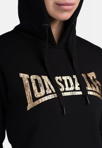 Black hoodie with a gold distressed logo "Lonsdale 15." Features a drawstring hood and ribbed cuffs. Soft fabric texture.