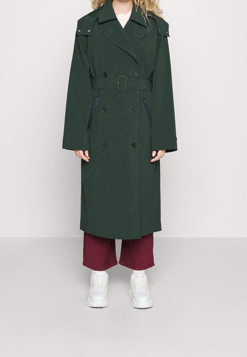 Personne portant un trench-coat vert foncé ceinturé, un pantalon large bordeaux et des baskets blanches épaisses, debout devant un fond clair uni.