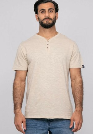 Homme avec cheveux noirs courts et barbe, portant une chemise henley beige à manches courtes et un jean bleu, debout devant un fond uni.