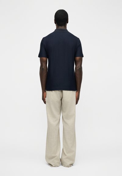 Άνδρας που στέκεται με την πλάτη προς τα εμπρός, φορώντας μπλε polo με κοντά μανίκια και ανοιχτό μπεζ παντελόνι με φαρδιά μπατζάκια, σε απλό φόντο.