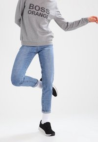 Sudadera gris con estampado "BOSS ORANGE", vaqueros azul claro y zapatillas negras. El conjunto presenta un ajuste relajado y vaqueros hasta el tobillo.