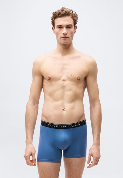 Νέος άνδρας όρθιος, φορώντας μπλε boxer briefs Polo Ralph Lauren με μαύρη μέση, ουδέτερη έκφραση, απλό ανοιχτό φόντο.