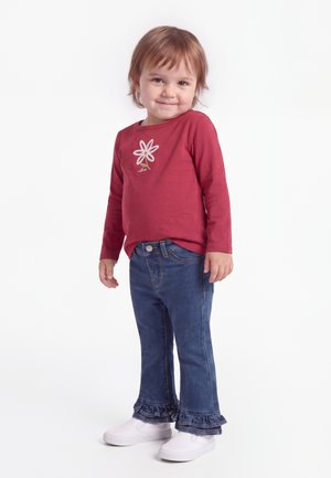 Tout-petit portant un t-shirt rouge à manches longues avec un imprimé floral, un jean bleu avec des volants à l'ourlet et des chaussures blanches à enfiler, debout sur un fond blanc.