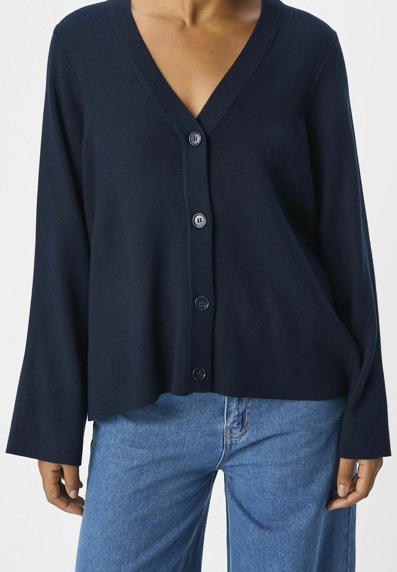 Femme portant un cardigan bleu marine à col en V boutonné avec des manches longues et un jean en denim bleu clair, debout devant un fond uni.