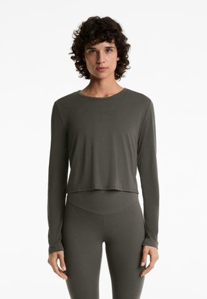 Vrouw met kort krullend haar, gekleed in een strakke lange mouw crop top en hoog getailleerde legging in dezelfde donkergrijze kleur.