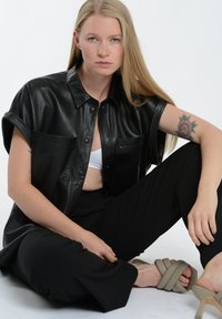 Sort læder kortærmet skjorte med to forlommer, parret med sorte bukser og beige strophæle, siddende positur, glatte teksturer.