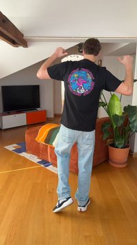 Man pointing thumbs at colorful graphic design on the back of his black t-shirt, standing in a living room with plants and furniture.