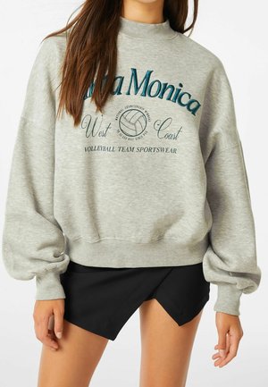 Vrouw die een grijze oversized sweatshirt draagt met de tekst "Santa Monica West Coast Volleyball Team Sportswear" en zwarte asymmetrische shorts.