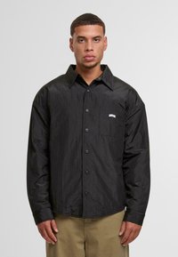 Fekete gombos ing, amely texturált anyagból készült, hegyes gallérral, hosszú ujjal és mellzsebbel rendelkezik, rajta "UNFAIR" logóval.