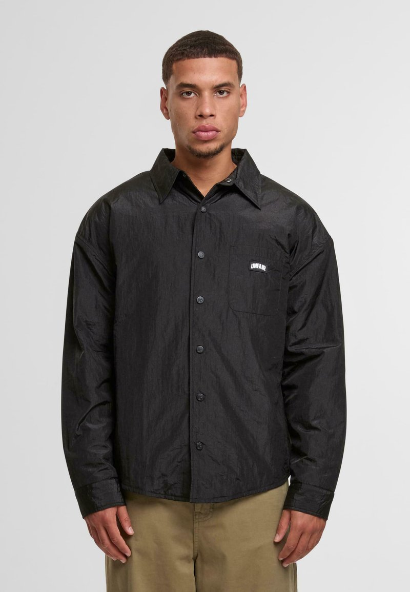 Fekete gombos ing, amely texturált anyagból készült, hegyes gallérral, hosszú ujjal és mellzsebbel rendelkezik, rajta "UNFAIR" logóval.