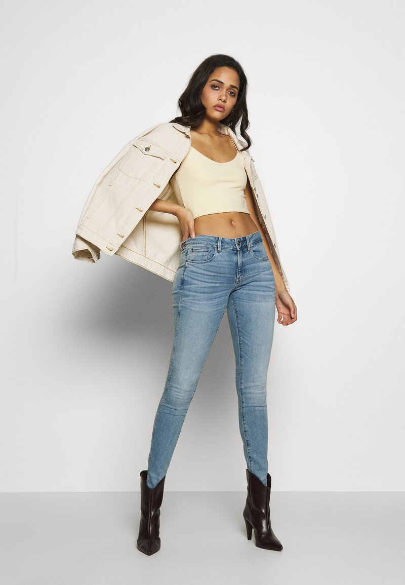 Un jean skinny bleu clair avec une texture délavée, associé à un top beige court et une veste beige. Les accessoires comprennent des bottes à talons noirs.