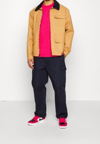 Tan jacket with a dark collar, pink t-shirt, navy cargo pants, and pink and black sneakers. Multiple pockets on the jacket and pants.