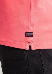 T-shirt en coton rose avec un éclat latéral inférieur, arborant une étiquette bleue portant la mention "Petrol Ind." et un jean noir contrastant en arrière-plan.