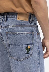 Pogled s leđa na osobu koja nosi svijetloplave traperice s vezenim kaktusom i suncem na stražnjem džepu te smeđu košulju.