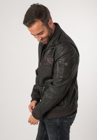 Schwarze Leder-Bomberjacke mit gerippten Bündchen und Saum, die mehrere Taschen und gestickte Patches auf der Brust und dem Ärmel aufweist.