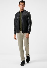 Schwarze Lederjacke mit einer Reißverschlusstasche auf der Brust, olivgrüner Rundhalsausschnitt-Pullover, hellbeige Hose und schwarze Stiefeletten.