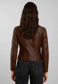 Giacca di pelle marrone con un design aderente, maniche lunghe e cuciture posteriori; abbinata a jeans neri a vita alta, capelli voluminosi in vista.