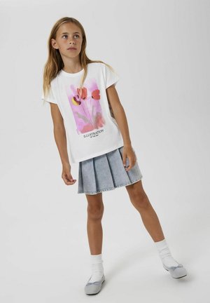 T-shirt blanc avec une illustration de fleurs roses et du texte noir. Associé à une jupe en denim plissée de couleur bleu clair, porté avec des chaussettes blanches et des chaussures grises.