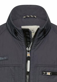 Donkergrijze jas met opstaande kraag, centrale ritssluiting gedeeltelijk dicht en een klein ritssluitingszakje op het rechterborstgedeelte.