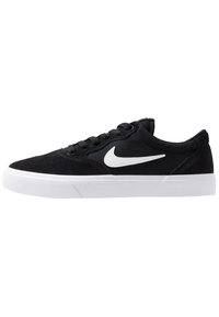 Černé semišové tenisky s bílými gumovými podrážkami, které mají na straně bílé logo Nike, kulatou špičku a klasický design na šněrování.
