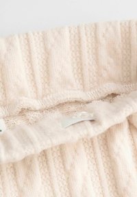 Jasnobeżowa dzianina o prążkowanej fakturze z małymi wzorami w kształcie rombów. Posiada falbaniaste wykończenie oraz wszytą metkę wewnątrz.