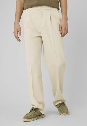 Homme portant un pantalon plissé beige avec des espadrilles vertes à lacets et un haut en tricot marron, debout devant un fond uni.