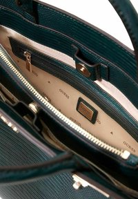 Sac à main texturé bleu sarcelle foncé avec fermeture éclair dorée, doublure intérieure crème et imprimé logo. Comprend une poche supplémentaire et des détails en métal poli.