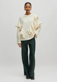 Crème kleurige trui met gelaagd ruffle ontwerp aan één kant, gecombineerd met donkergroene, glanzende, wijde leren broek en zwarte schoenen.