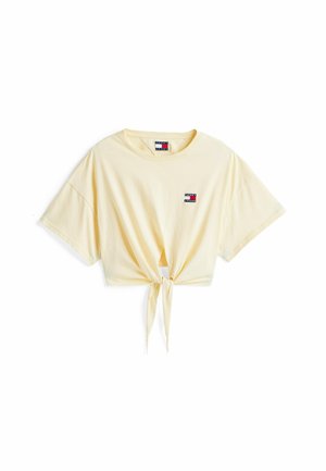 Camiseta amarilla corta, de algodón, con mangas cortas y un detalle de nudo en el frente. Presenta un pequeño parche de logo en colores rojo, blanco y azul.