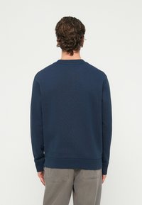 Muž s kučeravými hnedými vlasmi, oblečený v námornícky modrém tričku s dlhým rukávom a sivých nohaviciach, stojaci chrbtom k fotoaparátu.