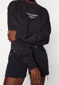 Svart sweatshirt med vit text "Calvin Klein Jeans", ribbat rundad halsringning och avslappnad passform, parat med svarta denimshorts med fransat avslut.