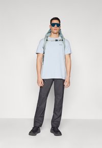 Light blue short-sleeve shirt, gray pants, black shoes, and a backpack with green accents. Reflective sunglasses enhance outdoor functionality.