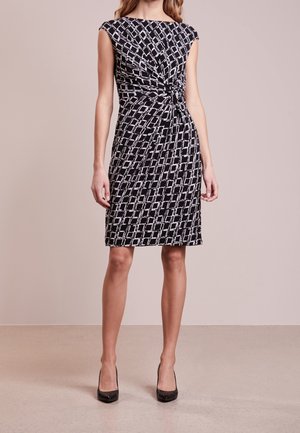 Czarna sukienka bez rękawów do kolan z białym geometrycznym wzorem i skręconym węzłem w talii, noszona z czarnymi szpilkami.