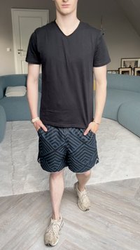 Černé tričko s výstřihem do V, vzorované černé a modré šortky a světle hnědé tenisky. V pozadí se nachází světlý koberec a měkká pohovka.